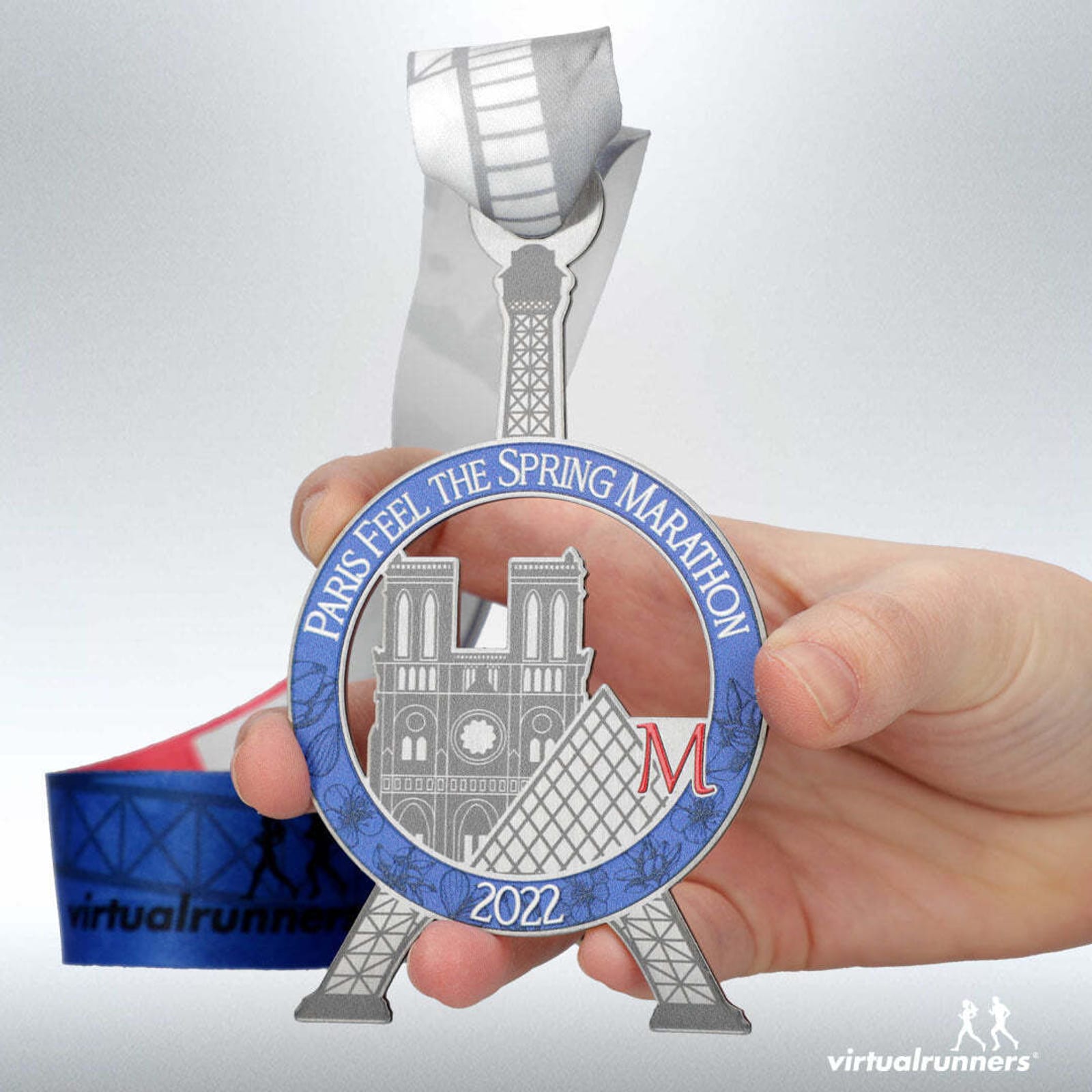 A hand holding a Paris-themed marathon medal with "PARIS FEEL THE SPRING MARATHON 2022" inscribed. The medal depicts the Eiffel Tower and Notre-Dame Cathedral. The ribbon attached has a gray and blue design with the logo "virtualrunners".