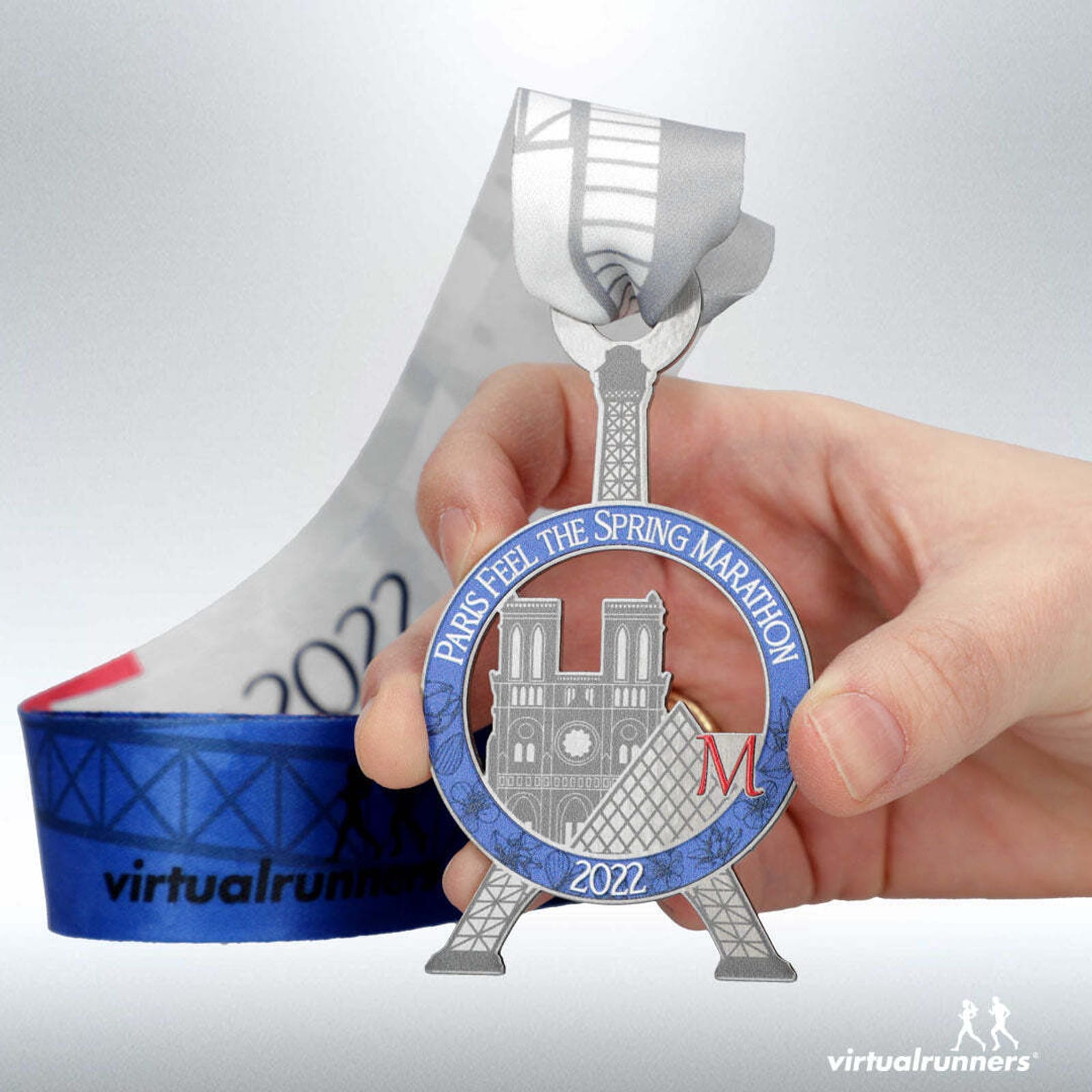 A hand holds a circular medal featuring the Eiffel Tower, Notre Dame, and the text "Paris Feel The Spring Marathon 2022." The ribbon attached to the medal has a blue and white gradient design with "2022" and "virtualrunners" printed on it.