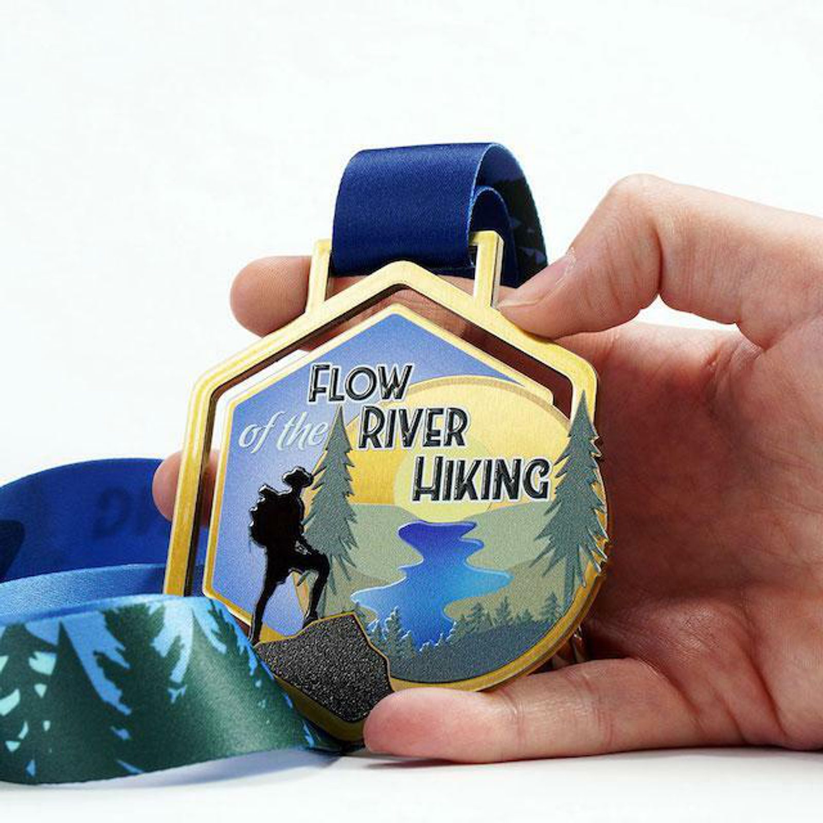 A hand holds a medal for "Flow of the River Hiking." The medal features an illustration of a hiker, pine trees, and a flowing river. It is attached to a blue ribbon decorated with tree designs.