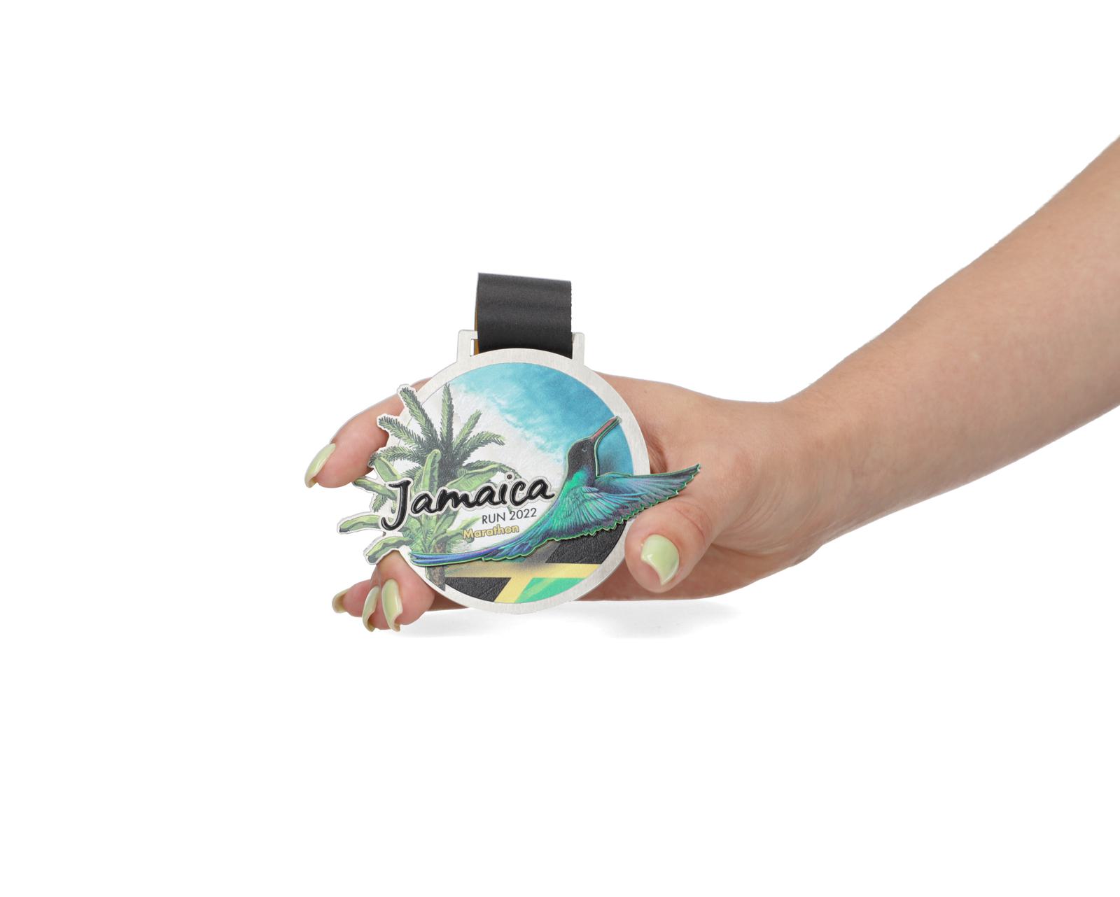 A hand holding a medal with the text "Jamaica Run 2022". The medal features a colorful design with a palm tree, a bird, and a scenic background that includes mountains and the sky.