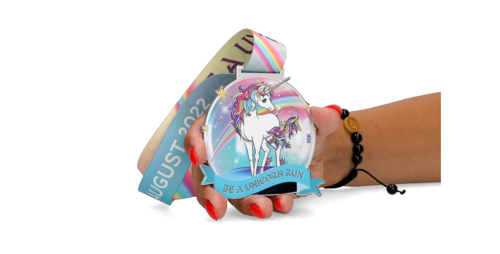 A hand with red nail polish holds a medal featuring a colorful unicorn with the text "BE A UNICORN RUN" on a banner. The ribbon attached to the medal has "AUGUST 2022" printed on it along with a rainbow design. A black bracelet adorns the wrist.