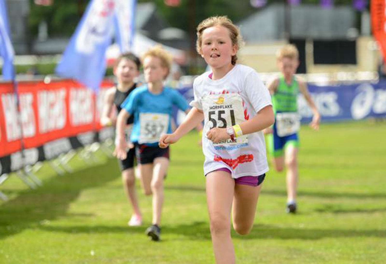 A young girl in a white shirt and purple shorts, wearing bib number 551, runs on a grassy field during a race. She is ahead of two other children in the background, who are also running and wearing bibs. Colorful flags and banners line the sides of the course.