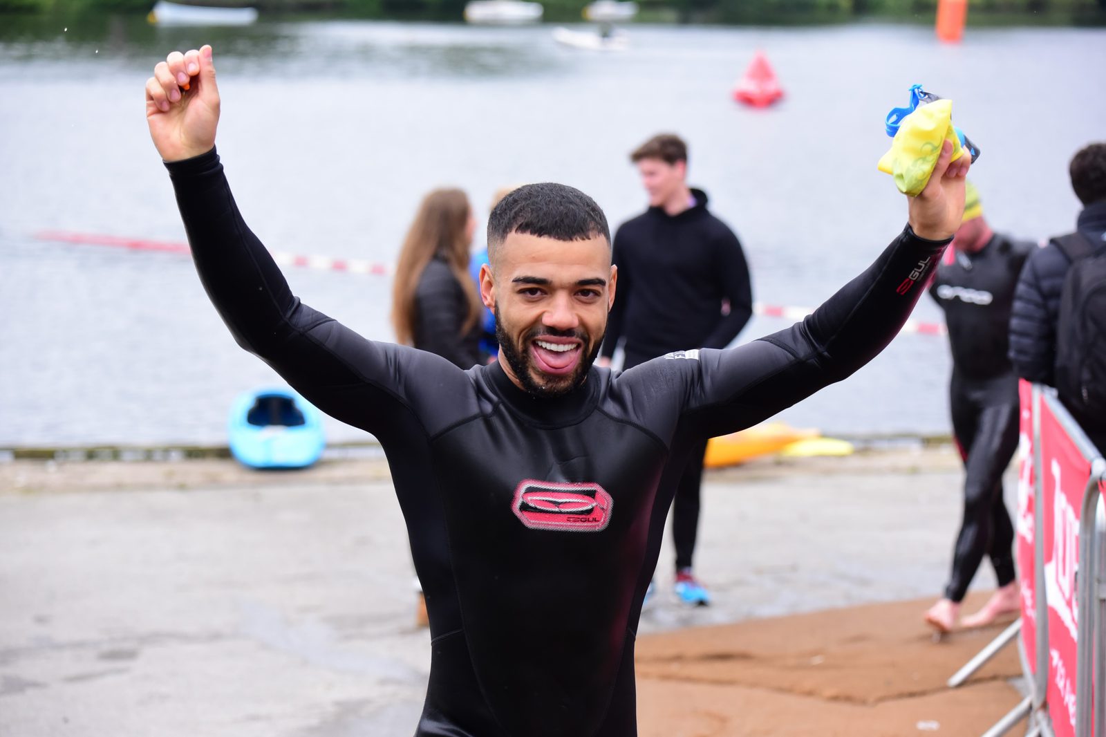 A man in a black wetsuit raises his arms in celebration after emerging from a lake, smiling broadly. Other swimmers and kayaks can be seen in the background, along with a crowd of spectators near the lake's edge.