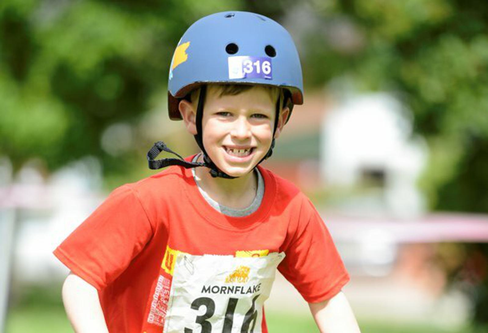 A young boy in a blue helmet and red shirt smiles while participating in an outdoor activity. He has a paper bib with the number 316 and the word “Mornflake” attached to his shirt. Trees and a blurred background suggest a park setting.