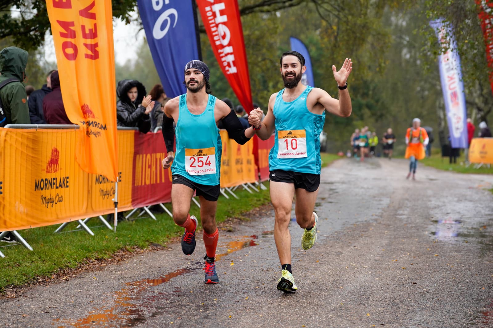 Two men wearing turquoise tank tops, black shorts, and race bibs 254 and 10, run hand in hand towards the finish line at an outdoor race event. Bystanders and colorful flags line the course. The man on the right is waving and smiling. The scene is wet and overcast.