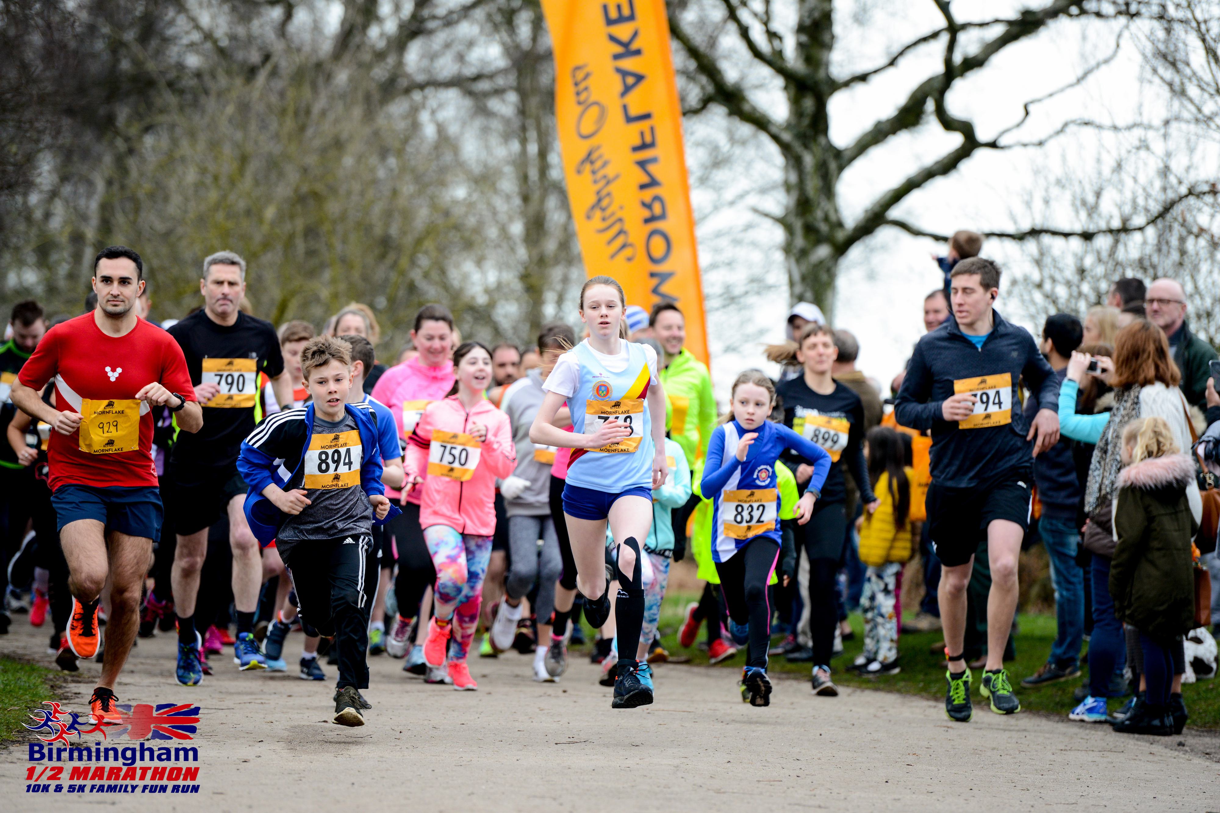 Runners, including children and adults, participate in the Birmingham 1/2 Marathon. They are seen in motion, wearing numbered bibs and athletic gear. A large orange banner and leafless trees are visible in the background on a cloudy day.