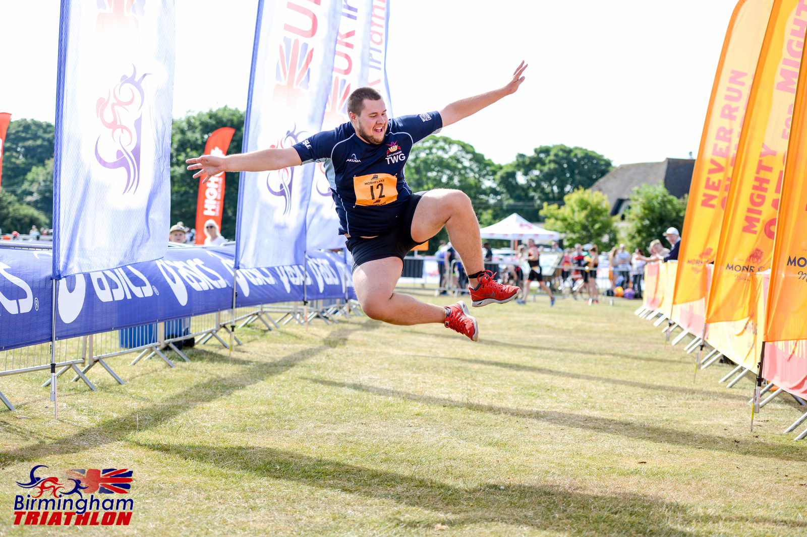 A participant in a Birmingham Triathlon is captured mid-jump near the finish line. He wears a black and blue athletic outfit with the number 12 visible on his chest. Various colorful banners and spectators can be seen in the background on a sunny day.