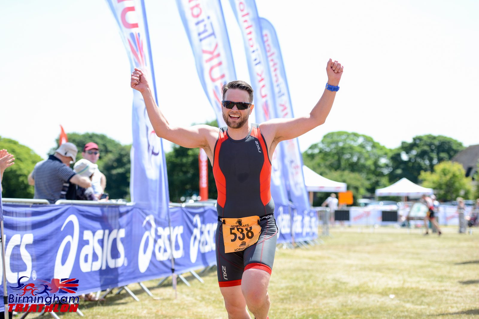 A man wearing a black and red triathlon suit and orange race bib number 598 raises his arms in celebration as he crosses the finish line at the Birmingham Triathlon. Background includes sponsor banners, other participants, and trees under a sunny sky.
