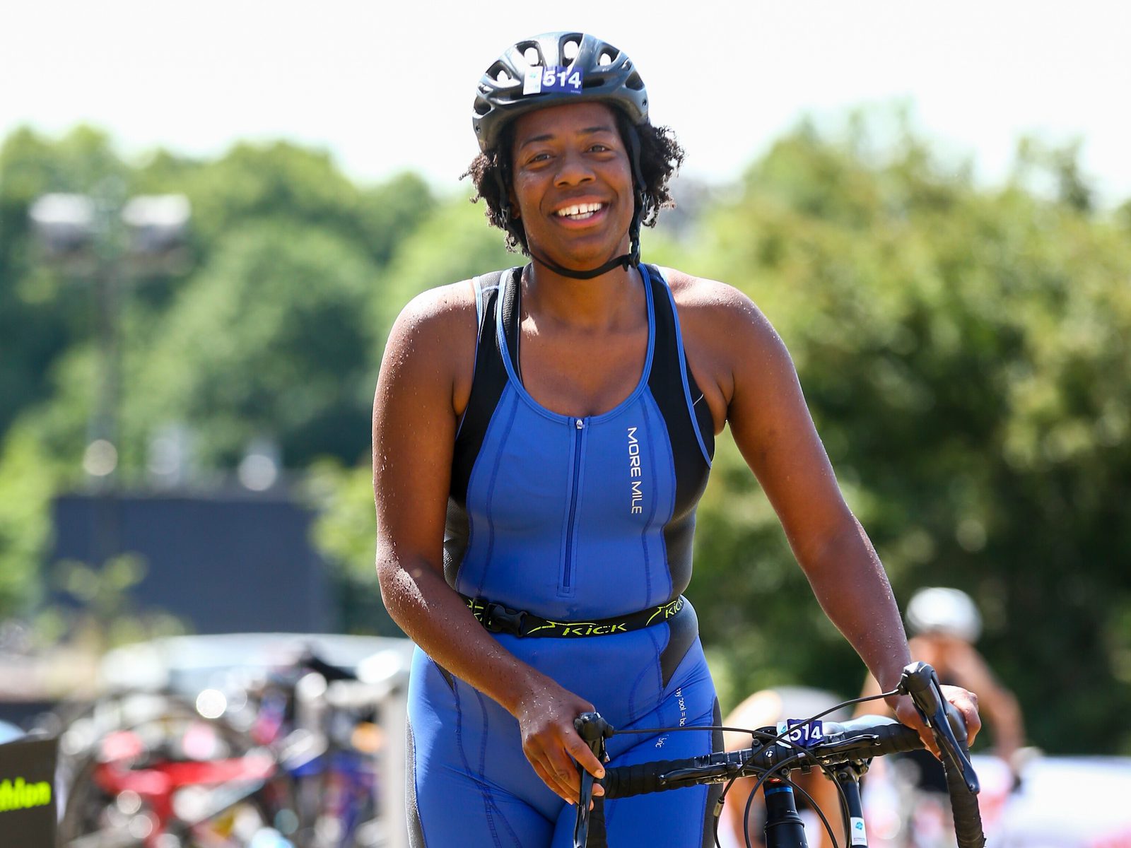 A smiling woman wearing a blue triathlon suit and black helmet stands with her bicycle. She appears happy and confident, with green trees and other cyclists in the background on a sunny day.