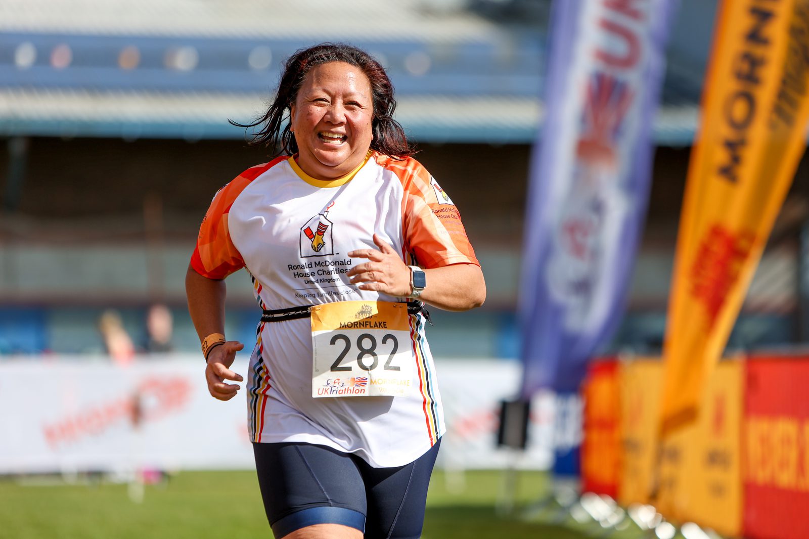A woman in a white and orange running shirt with the number 282 smiles as she crosses a marathon or race finish line. She has a race bib, and behind her are banners and structures indicating the event. The weather appears sunny and clear.