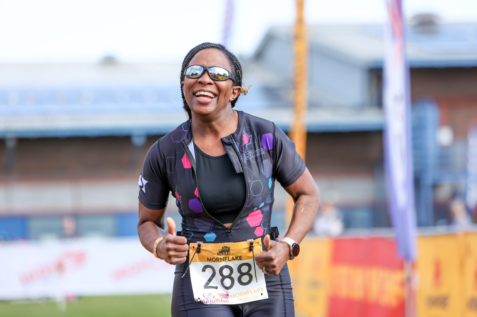 A woman wearing a cycling outfit and bib number 288 smiles while giving two thumbs up during a race. A banner and part of a building are visible in the background. She appears to be participating in a sporting event and has a joyful expression on her face.