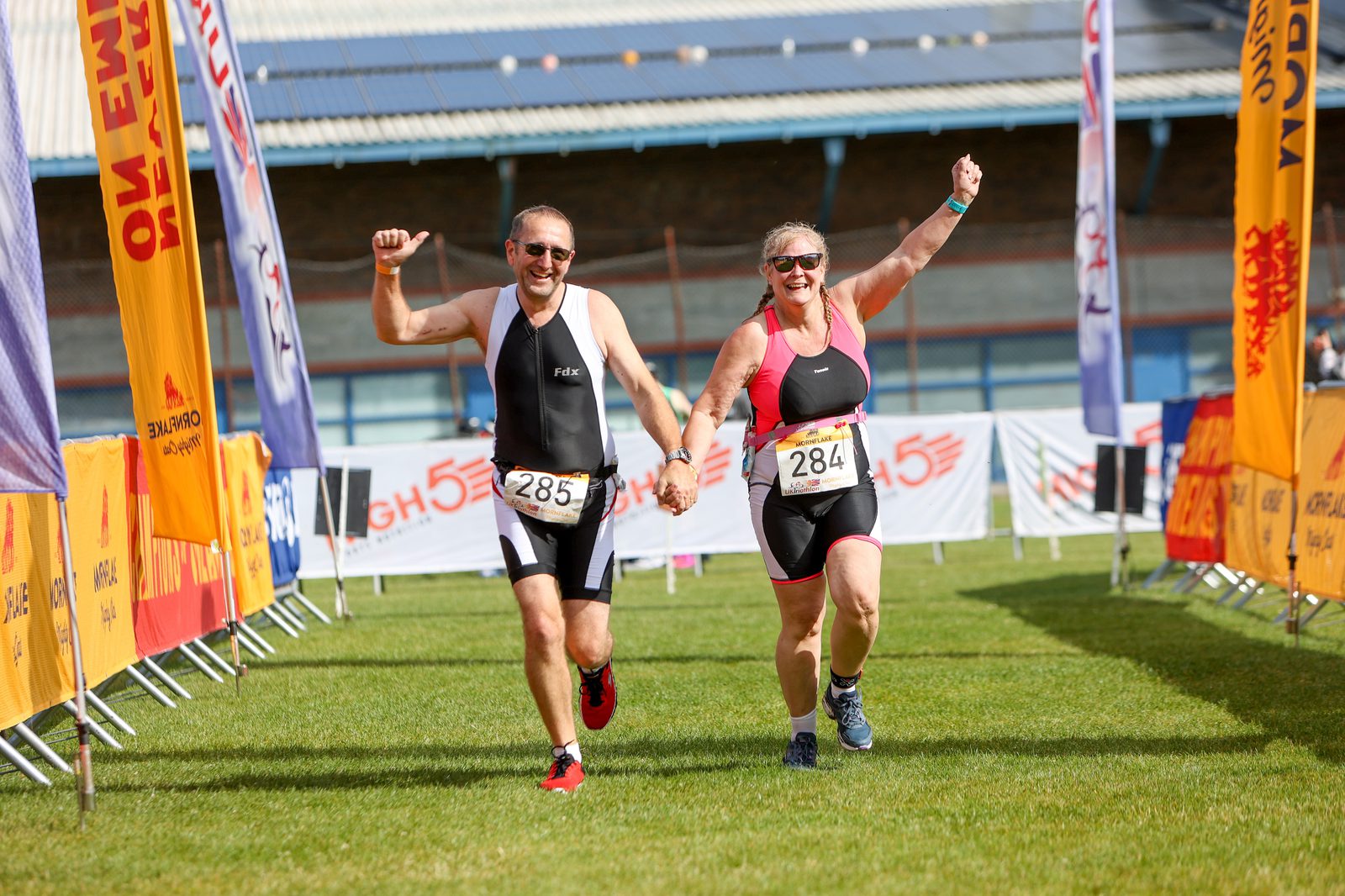 Two triathletes, a man and a woman, hold hands and cheer as they cross a finish line. Both are wearing black and red athletic gear with race numbers 285 and 284. Bright flags and a sunny outdoor setting frame their joyful moment.