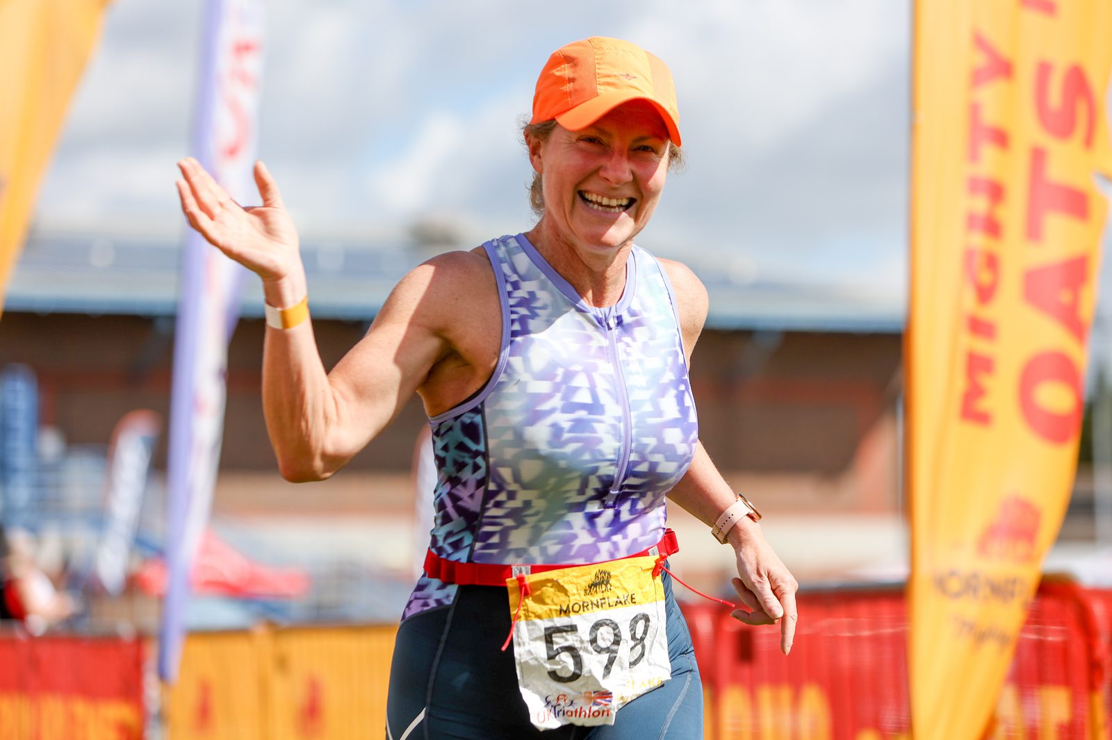 A woman wearing a blue and white tank top, orange cap, and race bib number 598 is smiling and waving at a race event. She is running past bright orange banners in the background that read "MIGHTY OATS." She appears happy and energetic.