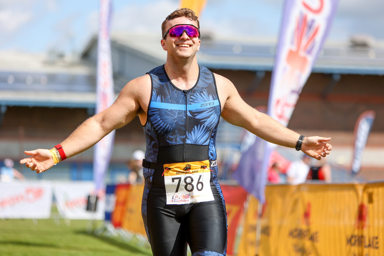 A male triathlete, wearing a black and blue tri suit and mirrored sunglasses, joyfully crosses the finish line of a race. His arms are outstretched, and he has a race bib with the number 786. Spectators can be seen in the background near banners and barriers.