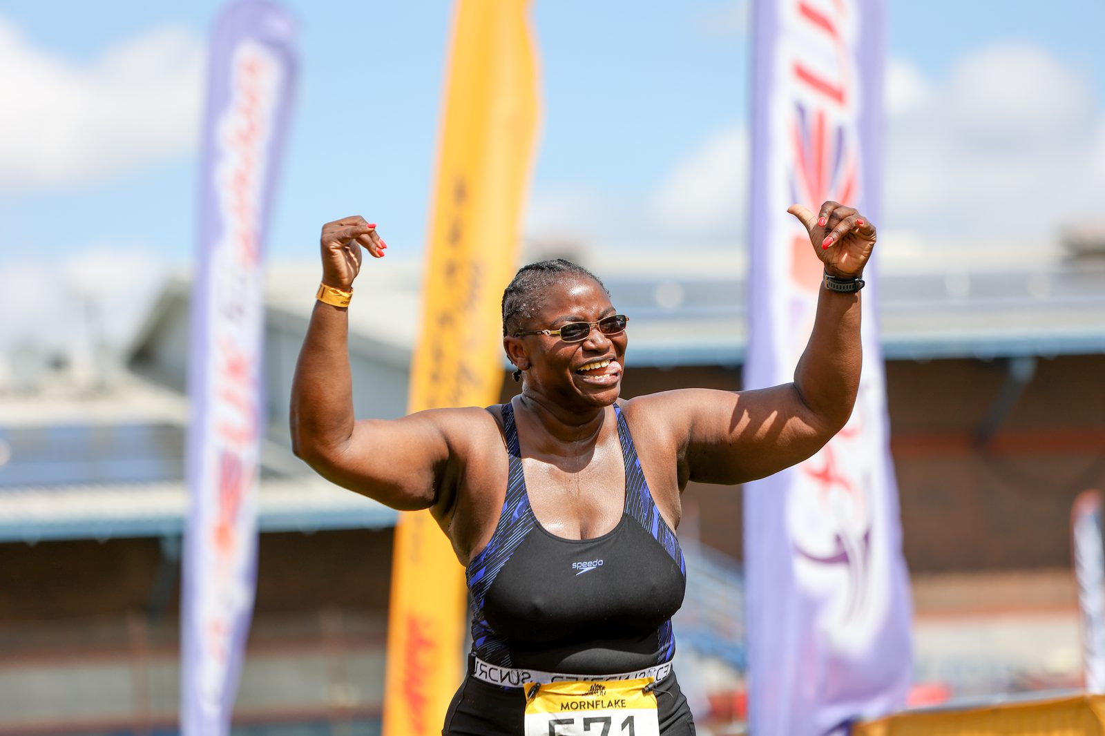A woman wearing a sports tank top and bib number 71 is smiling and raising her fists triumphantly. She is outdoors with a clear sky and sports banners in the background.