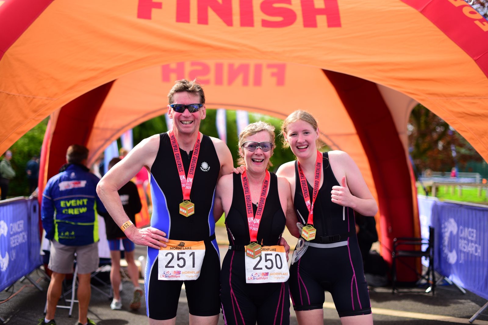 Three triathletes with race bibs 250 and 251 smile and pose for a photo under an orange "FINISH" arch. They wear medals around their necks and sporty outfits. The event crew and other participants are visible in the background.