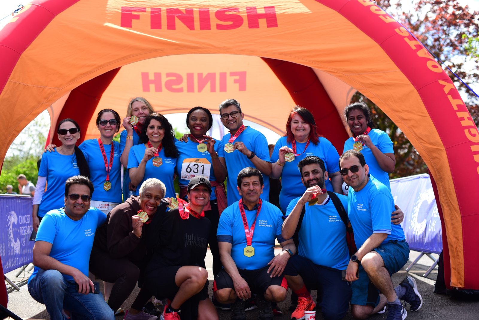 A group of people wearing blue shirts pose under an orange "FINISH" arch. They are smiling and holding up medals. A person in the middle kneels down holding a race bib numbered 631. The background includes trees and parts of a crowd barrier.