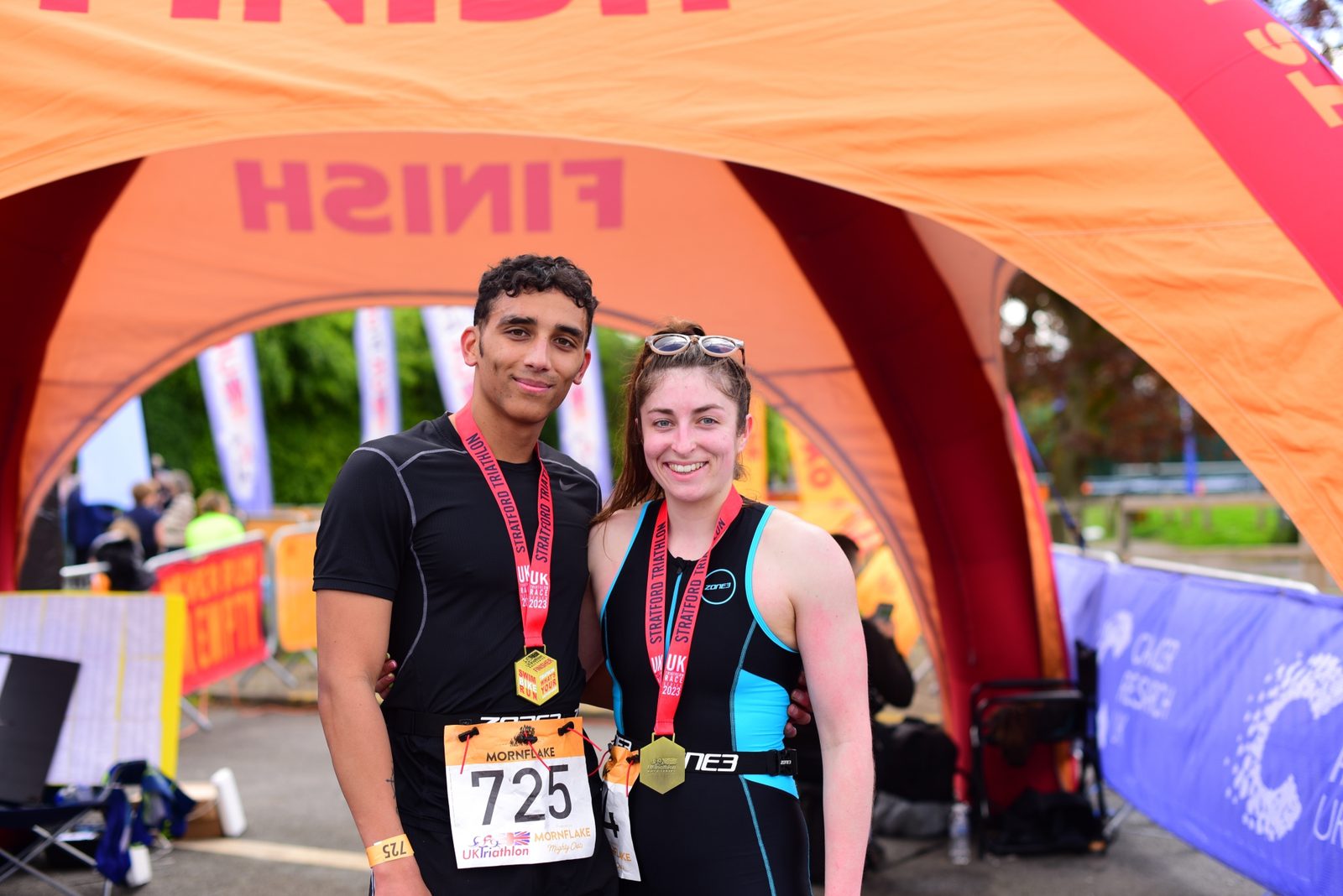 Two smiling athletes pose under an orange finish line arch after completing a race. One man on the left is wearing athletic clothes and a bib number 725, while the woman on the right is in a triathlon suit. Both have medals around their necks.