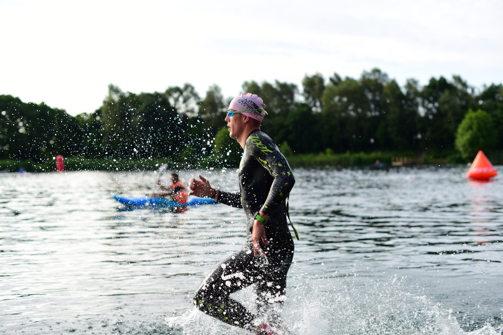 A person wearing a swim cap, goggles, and a wetsuit is emerging from a body of water during a triathlon. They are surrounded by splashing water with trees and kayakers in the background. An orange buoy is visible in the water.