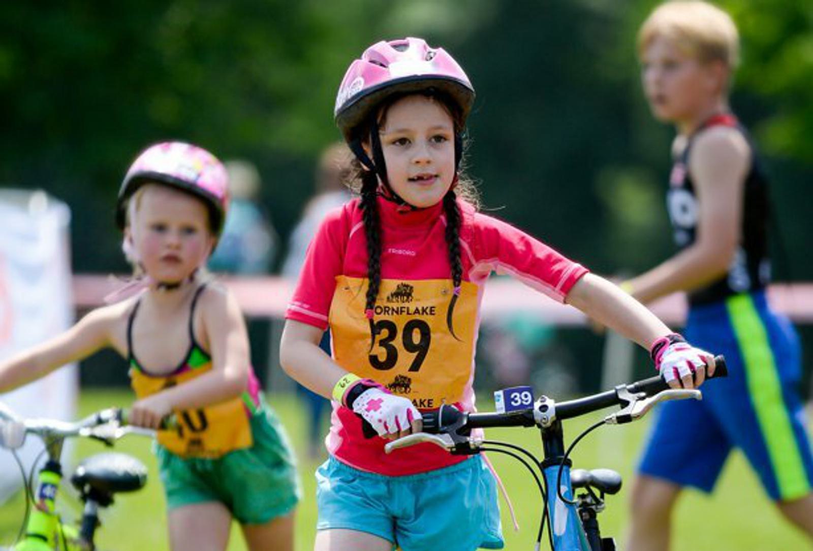 Three children wearing helmets and sporting biking attire are riding their bicycles outdoors. The child in front, with braided hair and wearing a red shirt and blue shorts, is marked with the number 39. The other two children are in the background, also on bikes.