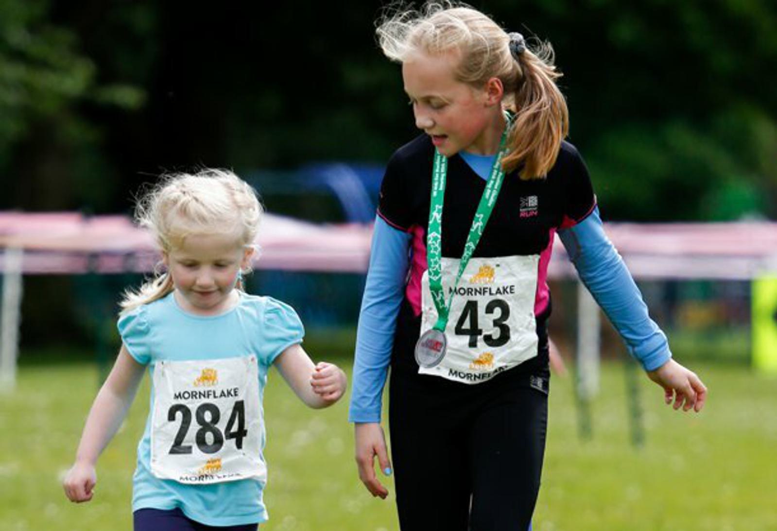 Two young girls with running bibs, numbered 284 and 43, walk together at an outdoor event. The older girl, wearing a medal, encourages the younger girl, who is looking down while walking. Both are dressed in athletic wear and appear happy and engaged.