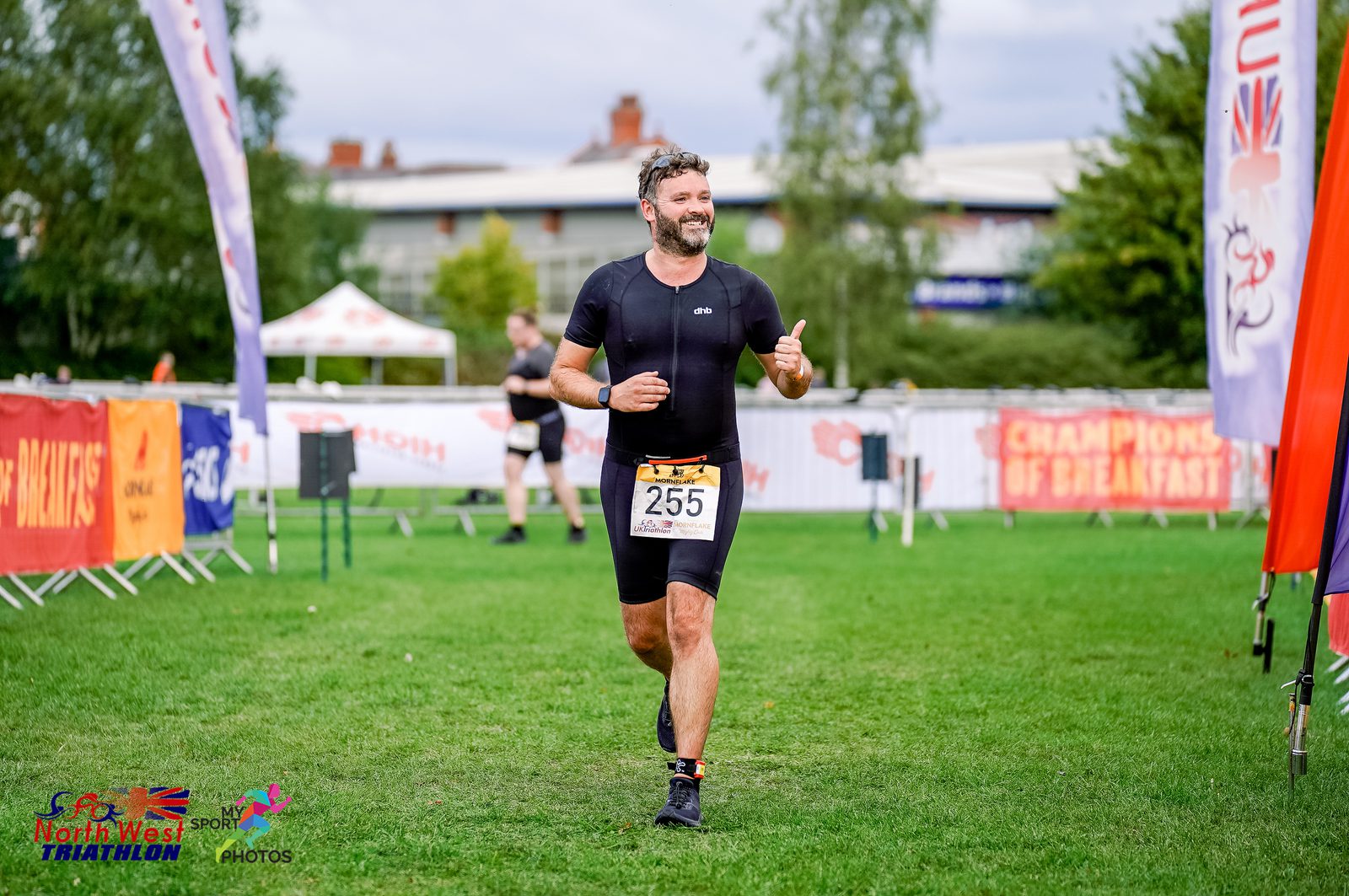 A man in black athletic wear and bib number 255 is running and smiling towards the finish line at the North West Triathlon. Banners, other participants, and supporters are visible in the background. The event is set in a grassy area with trees and buildings nearby.