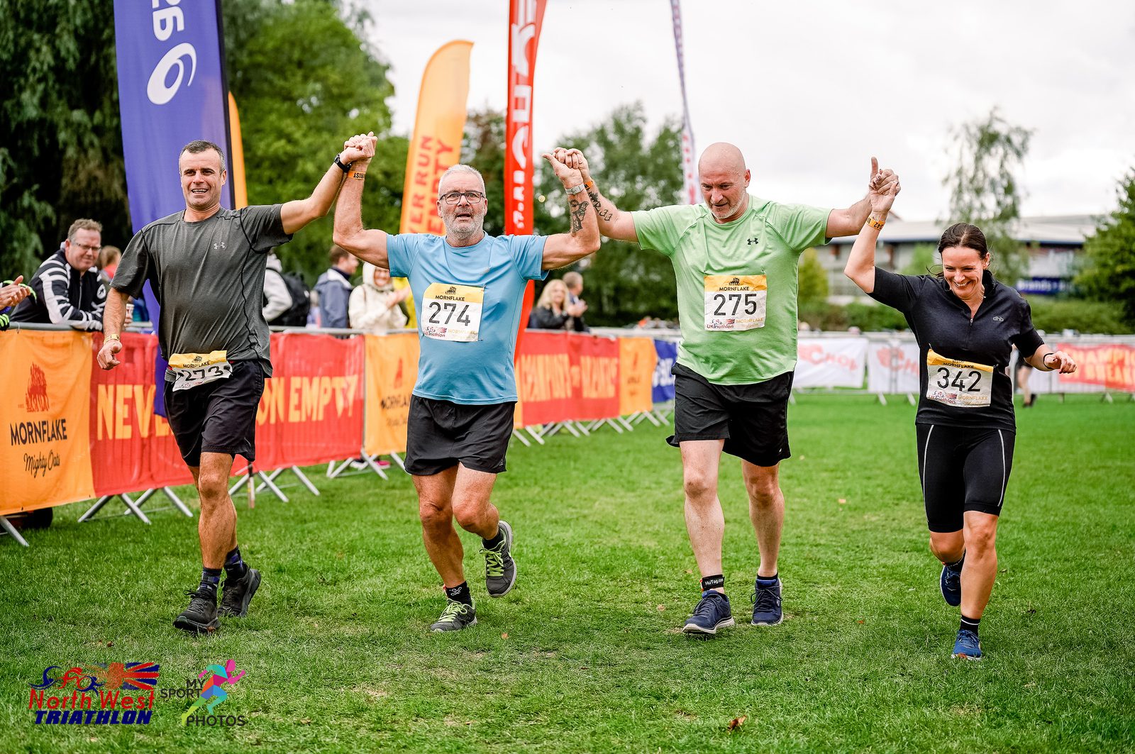 Four runners cross the finish line of a triathlon, holding hands and celebrating. They wear race bibs numbered 71, 274, 275, and 342. The background features cheering spectators, event banners, and flags for the Hever Castle Triathlon.