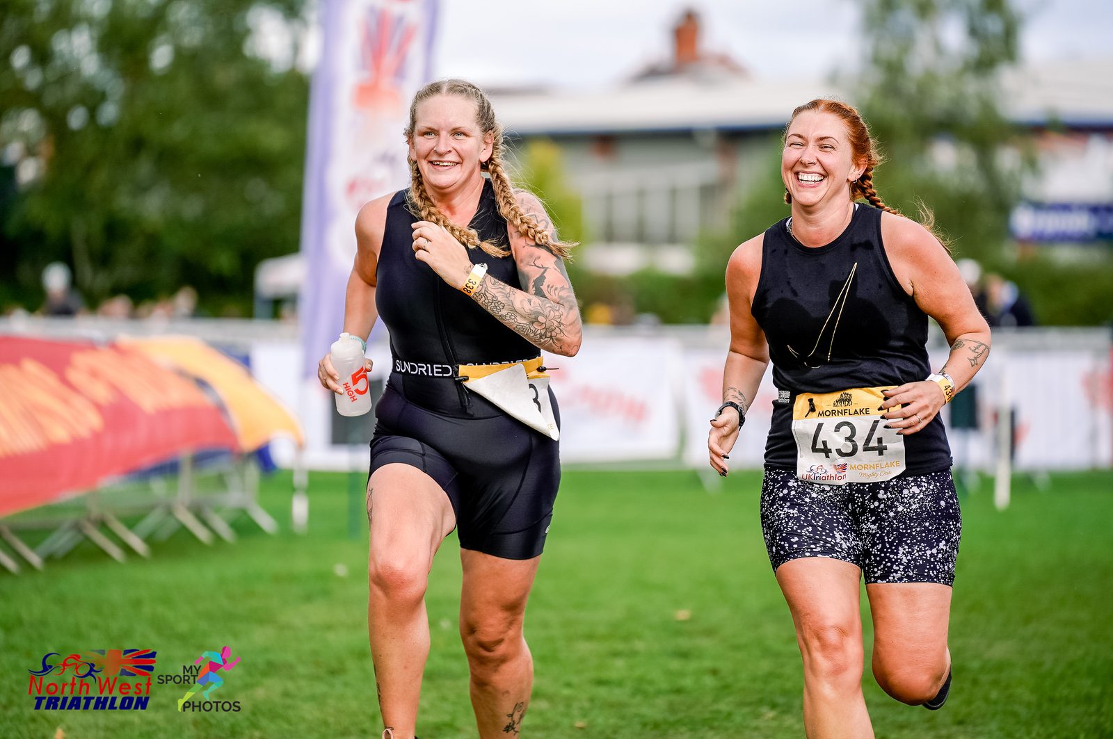 Two women, wearing athletic gear, are smiling and running towards the finish line of the North West Triathlon. They each have a race bib with numbers pinned to their outfits. A banner and a building are visible in the background.