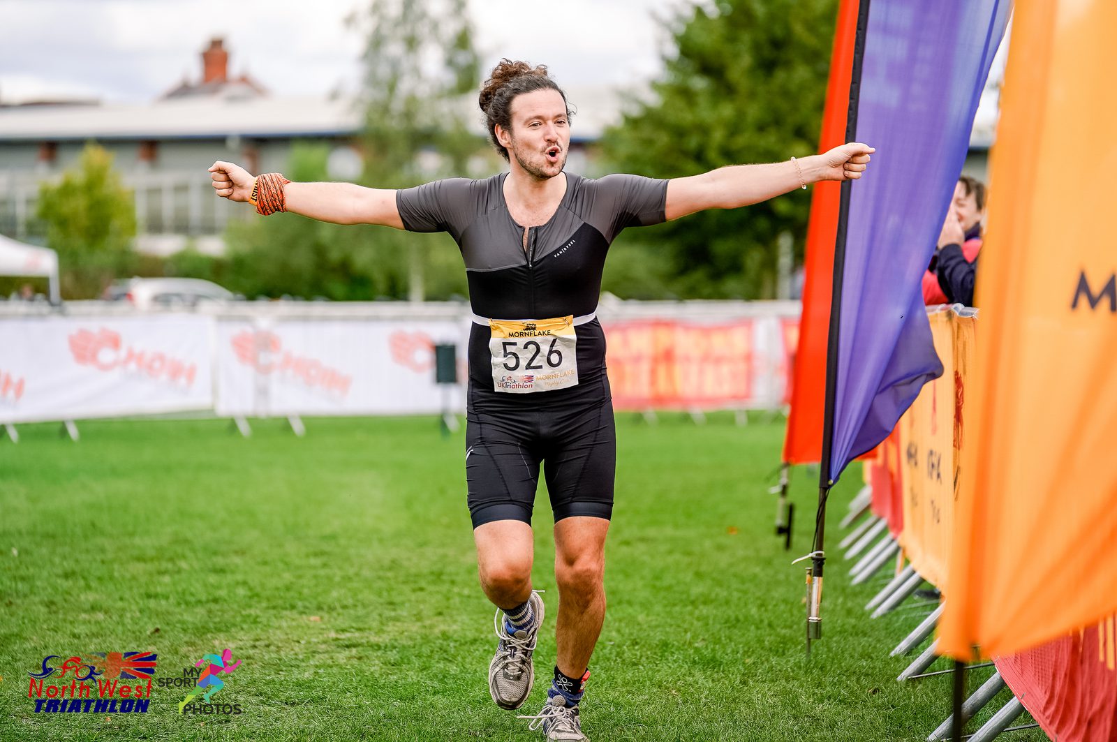 A man with bib number 526, wearing a black athletic outfit, is crossing the finish line of a race, arms outstretched in celebration. Colorful sponsor banners line the path. The event appears to be the North West Triathlon.