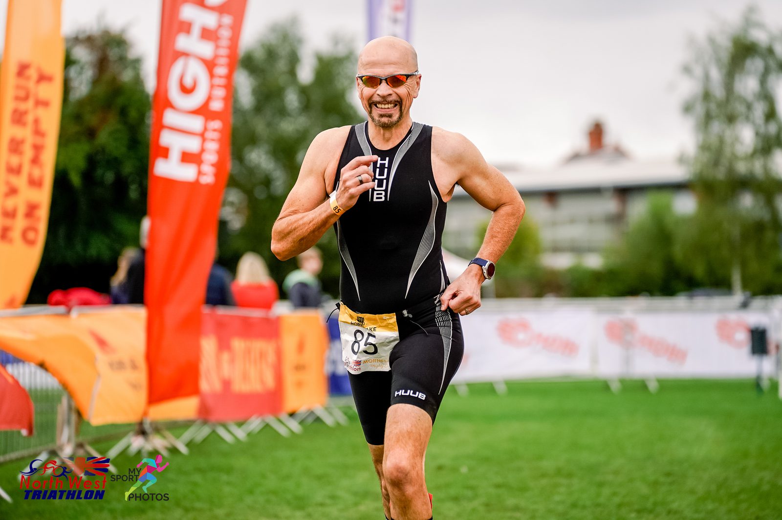 A man wearing sunglasses and a black triathlon suit runs with a determined expression during a triathlon event. He has a race number 85 on his waist. Spectators and colorful banners are visible in the background, including ones for HIGH5 and North West Triathlon.