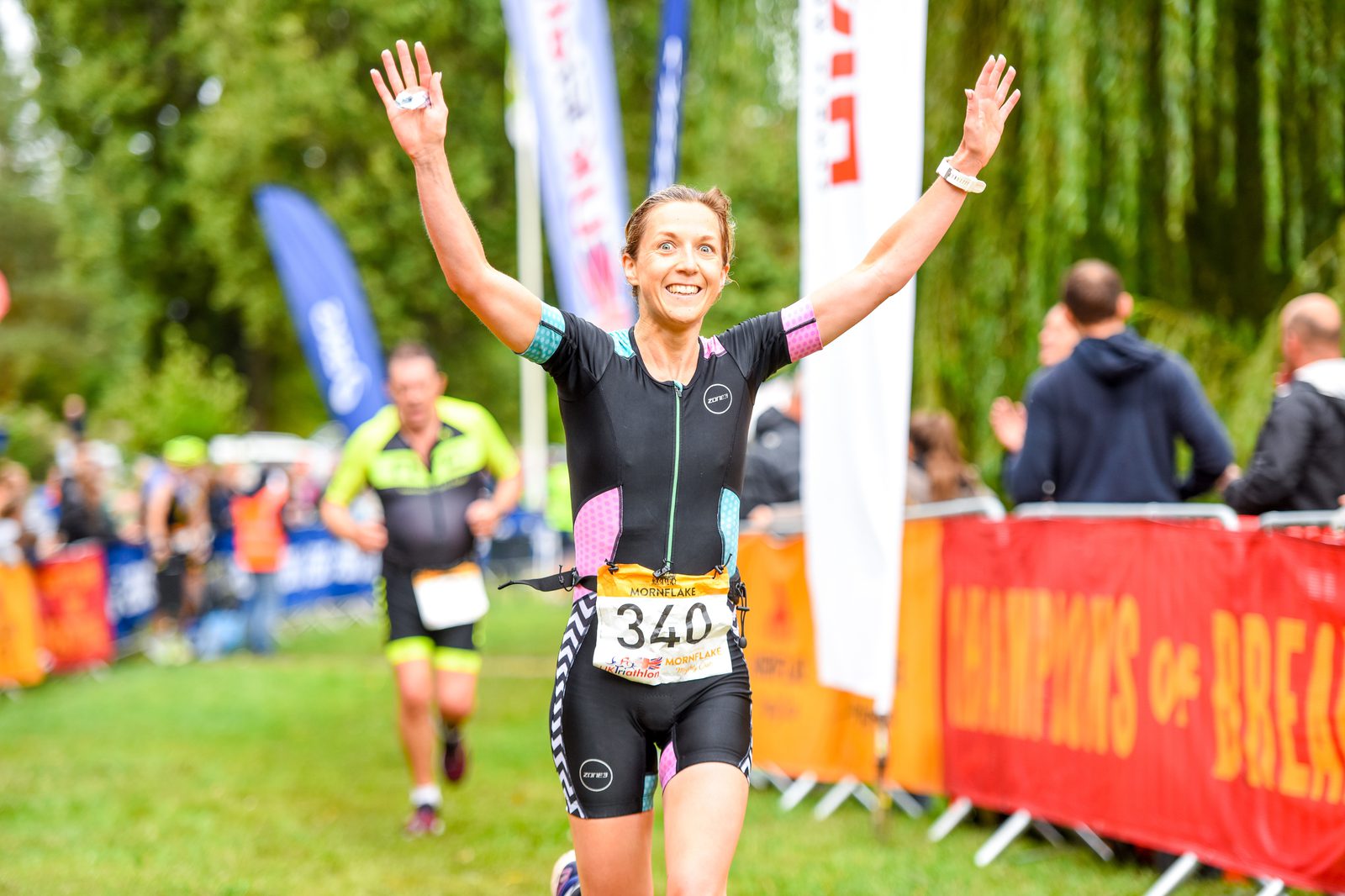 A female athlete, wearing a race number 340, crosses the finish line of a race with her arms raised in celebration. She's smiling, with other participants and banners visible in the background. The event appears to be outdoors with a grassy area and trees.