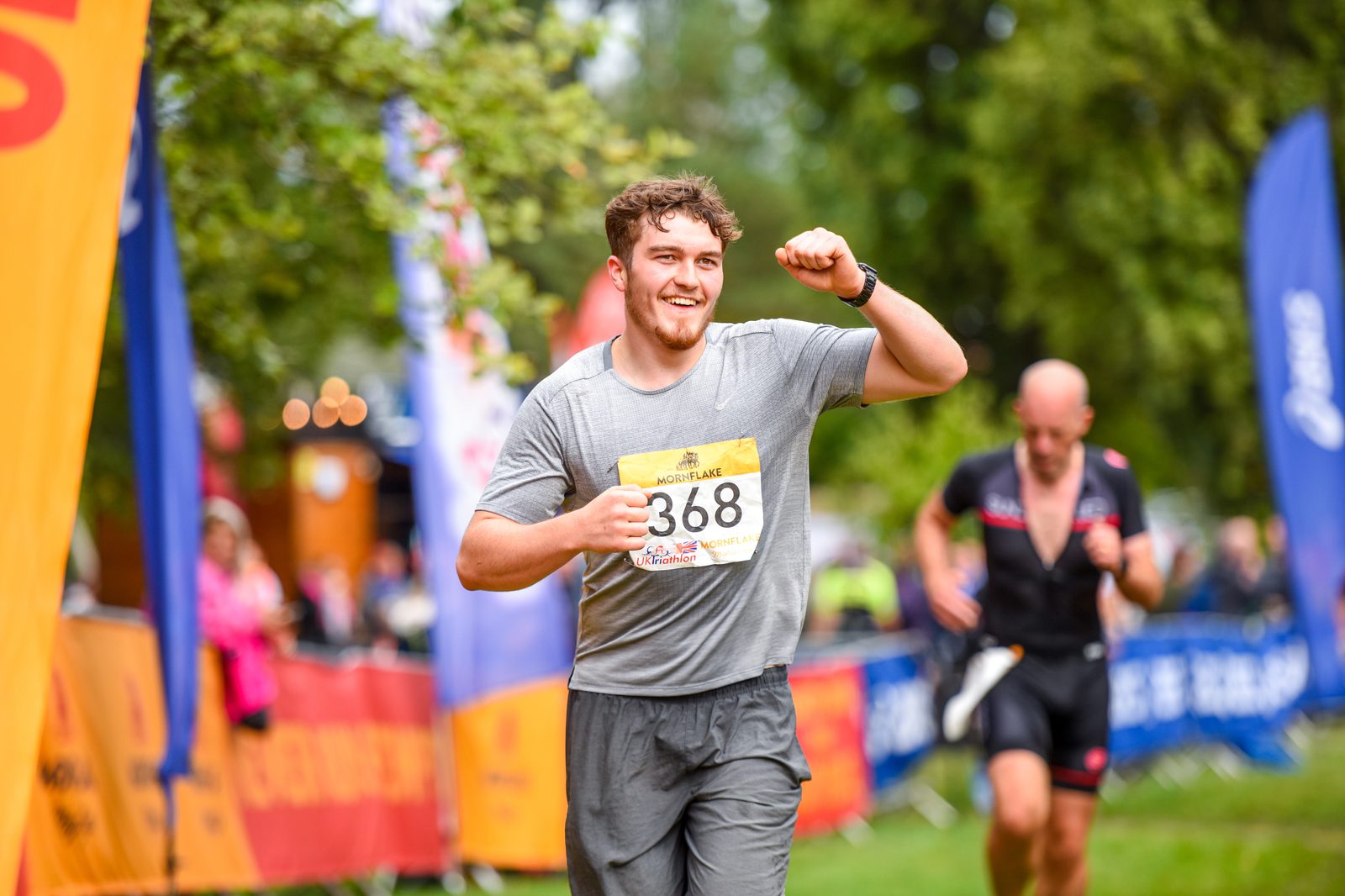 A runner wearing a gray shirt and bib number 368 raises his fist in celebration as he crosses a race finish line outdoors. People are seen in the background along the course, and colorful banners and flags are visible on either side.