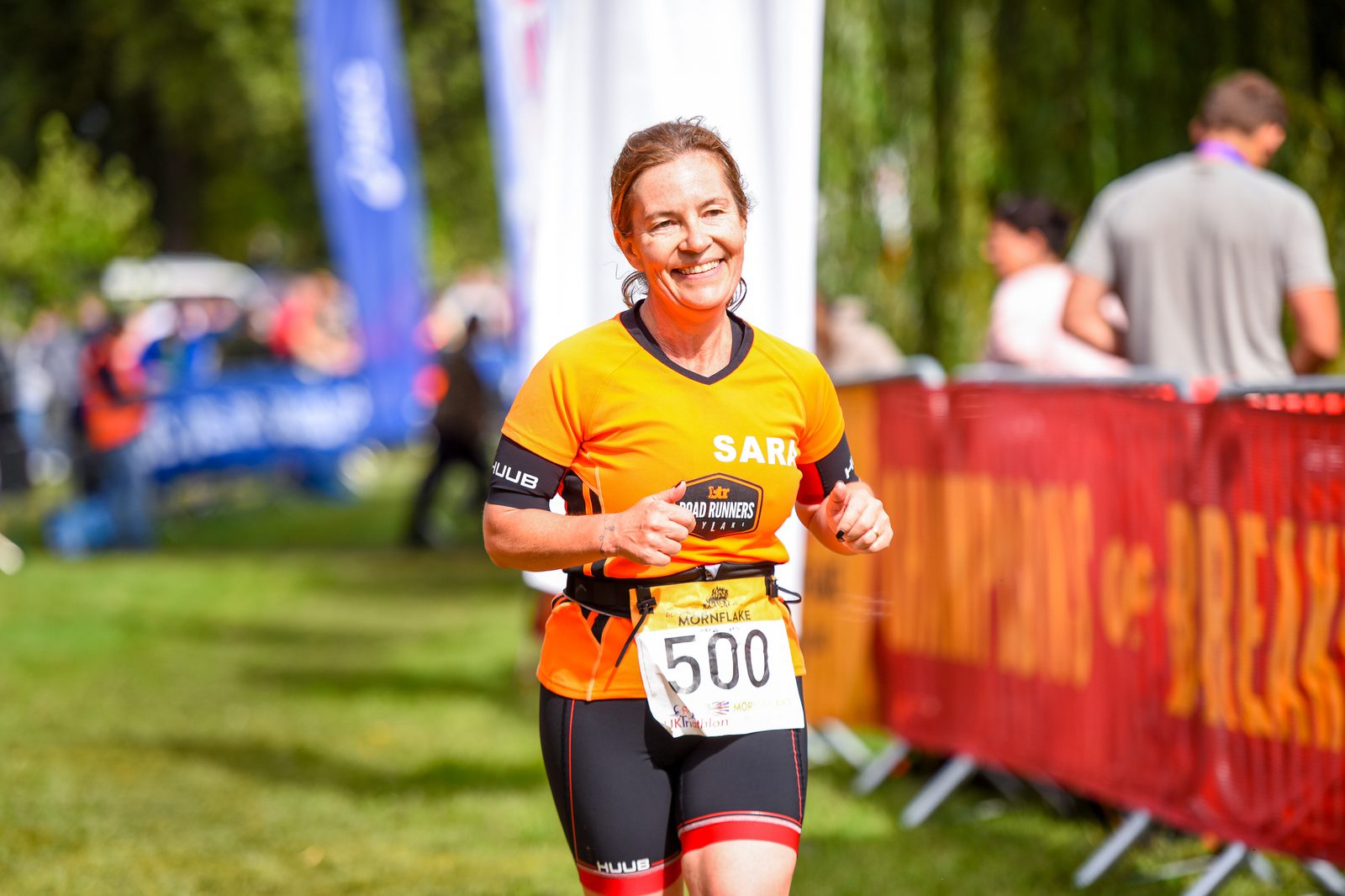 A woman wearing an orange and black athletic outfit with the number 500 bib is running in an outdoor event. She has a smile on her face and there are blurred spectators and banners in the background. The event seems to be taking place on a sunny day in a grassy area.