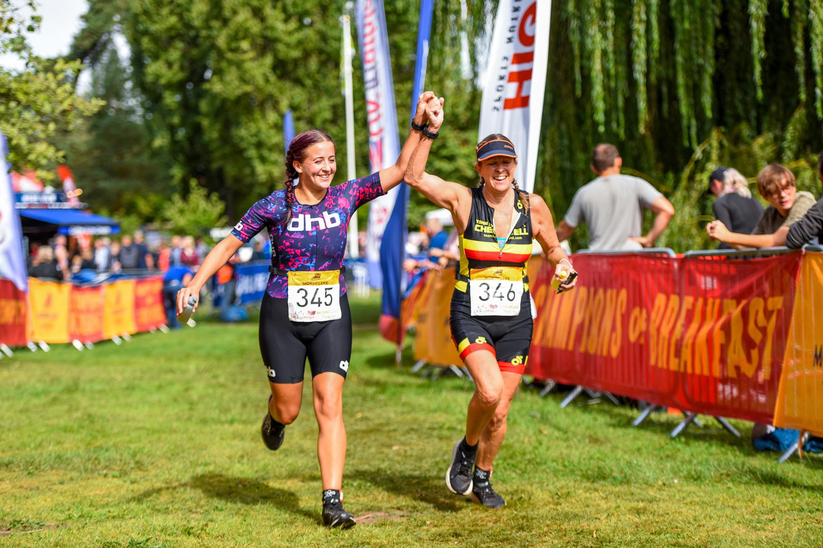 Two triumphant athletes cross the finish line of a race, holding hands and smiling. They wear numbered bibs 345 and 346, and colorful athletic wear. Spectators cheer behind barriers adorned with banners. Trees and lush greenery form the backdrop.