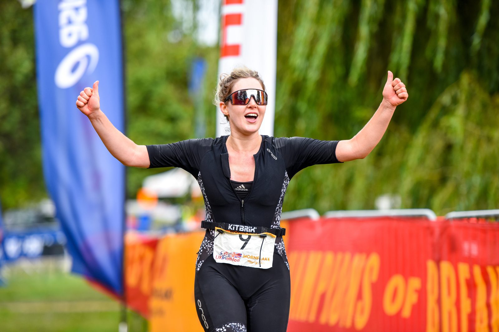 A triathlete crosses the finish line with a triumphant expression, raising both thumbs up. She is wearing sports sunglasses, a black racing suit, and a bib number. Colorful banners and spectators in the background suggest a lively race event.