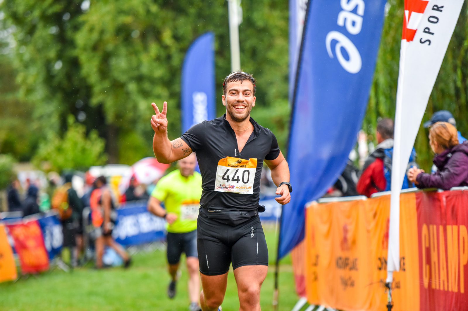 A smiling male athlete in black sportswear and race bib number 440 runs on a grassy path, holding up a peace sign with one hand. Colorful flags and banners surround the path, while other participants and spectators are visible in the background.
