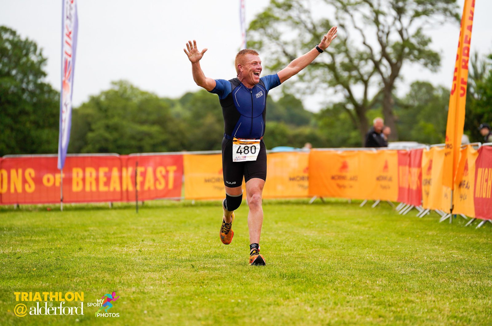 A triathlete wearing a blue and black wetsuit celebrates joyfully with arms raised while crossing the finish line at the Alderford Triathlon. He is wearing a race bib with the number 480. Colorful banners and flags line the finish area on a bright, grassy field.