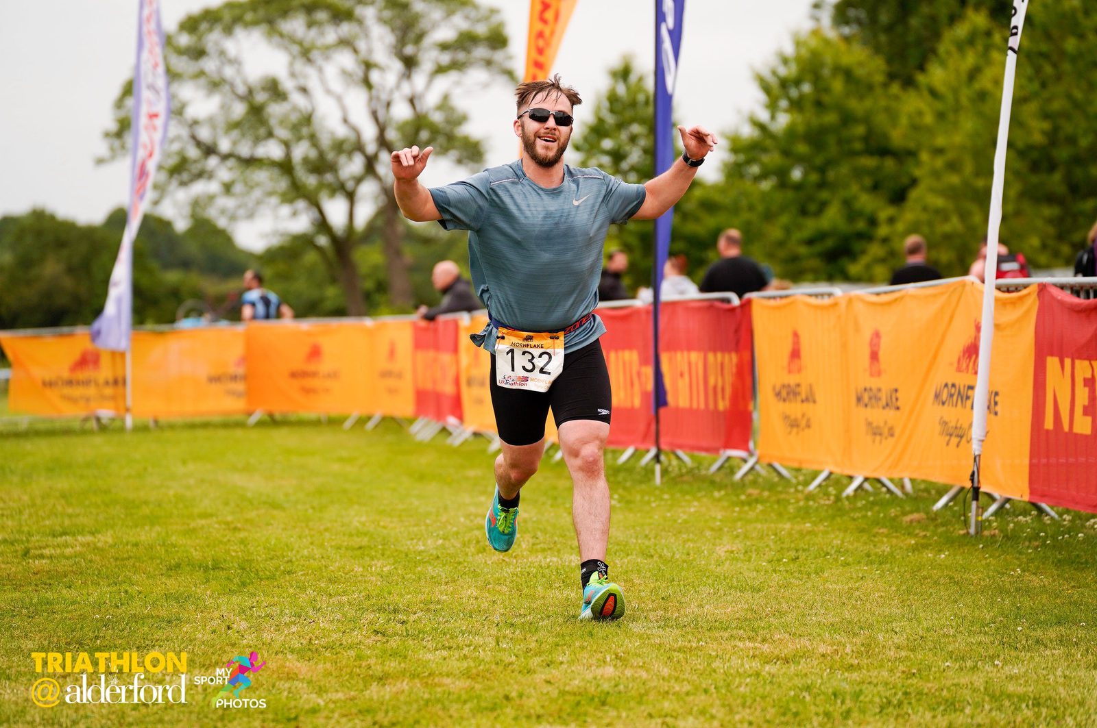 A man with a beard, wearing sunglasses, a blue shirt, running shorts, and athletic shoes, is joyfully running towards the finish line of a triathlon. He has his arms up in celebration, with bib number 132. Yellow and red banners line the grassy path.