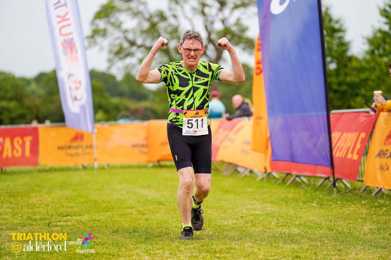 A man wearing glasses, a green and black shirt, and black shorts is finishing a race with his arms raised triumphantly. He has a race bib numbered 511. Colorful banners and spectators can be seen in the background. The setting appears to be an outdoor triathlon event.