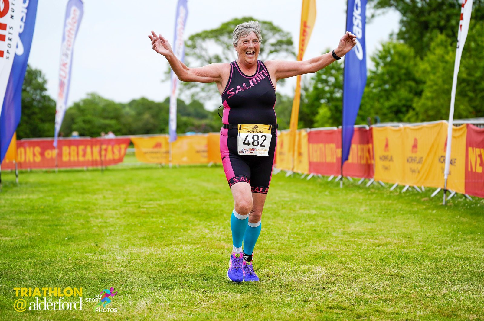 An older woman, wearing a black and pink triathlon suit, blue calf sleeves, and purple sneakers, crosses the finish line of a triathlon at Alderford. She has her arms raised in celebration, smiling broadly. Colorful banners and flags line the grassy path.