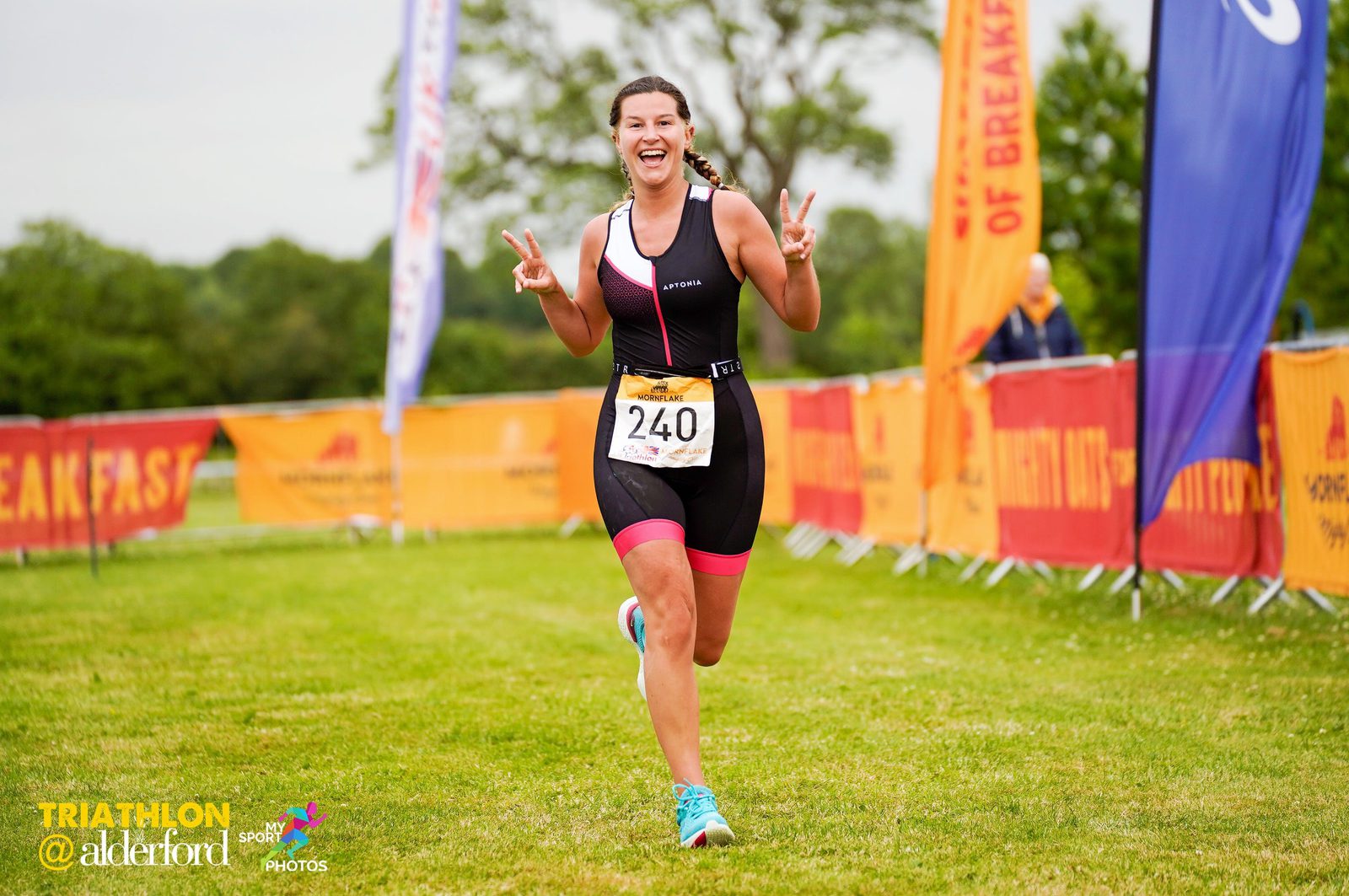A smiling woman wearing a black and pink triathlon outfit runs towards the camera, flashing a peace sign with both hands. She has a bib number 240 pinned to her front. Behind her are colorful banners and a grassy area, indicating the Triathlon @ Alderford event.