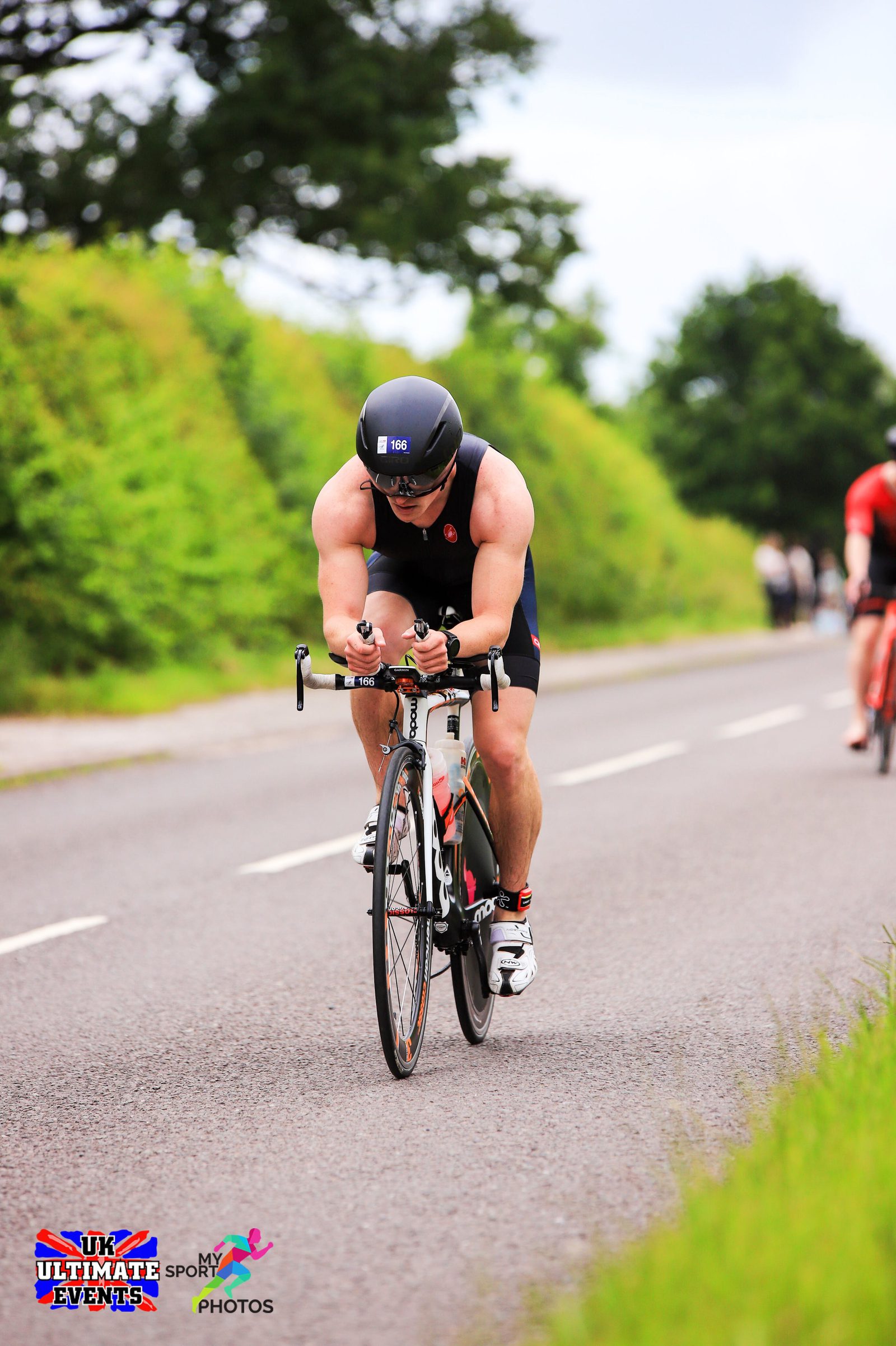 A cyclist wearing a helmet, sunglasses, and cycling gear, is riding on a road surrounded by greenery. Other cyclists are visible in the background. The event is marked by "UK Ultimate Events" and "My Sport Photos" logos at the bottom left corner of the image.