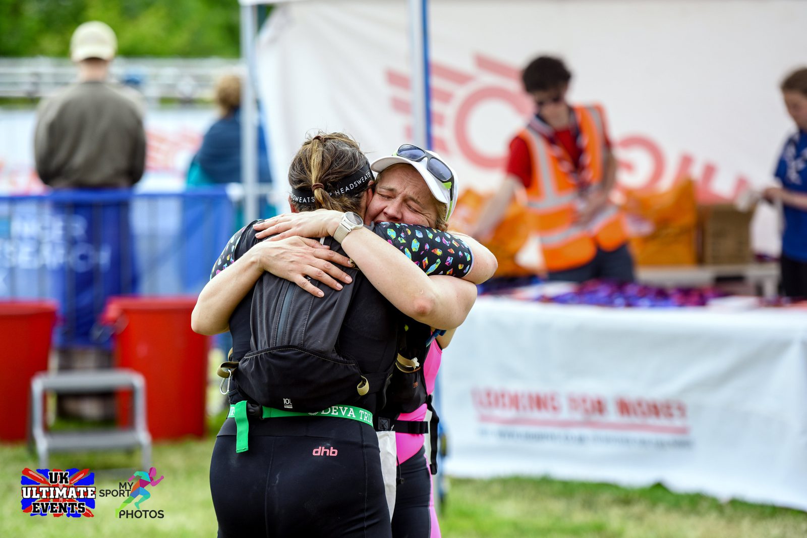 Two women, one wearing a black cap and the other in a white cap, share an emotional embrace at the finish line of a sporting event. A race volunteer stands in the background near a table with medals. Signs for "UK Ultimate Events" and "My Sport" are visible.