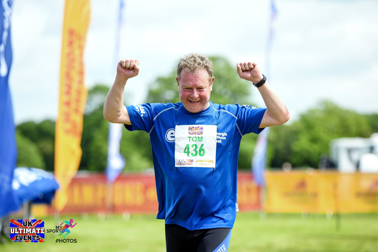 A man wearing a blue shirt and bib number 436 raises his arms in celebration as he crosses the finish line at an outdoor race event. The backdrop includes colorful banners, green trees, and a bright, clear sky. The event appears organized by UK Ultimate Events.