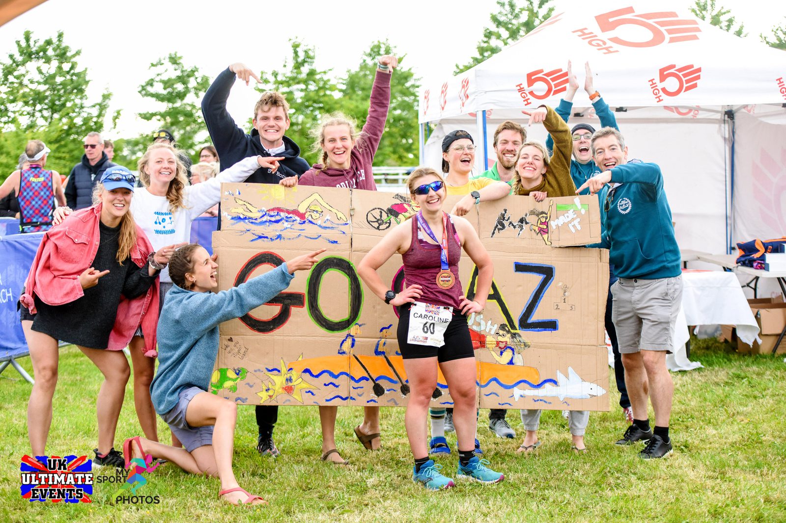 A runner, wearing a medal and race number 60, poses with a group of cheering supporters holding a large, decorated "GO AZ" sign at a race event. The supporters are smiling and gesturing triumphantly, with a tent and trees in the background.