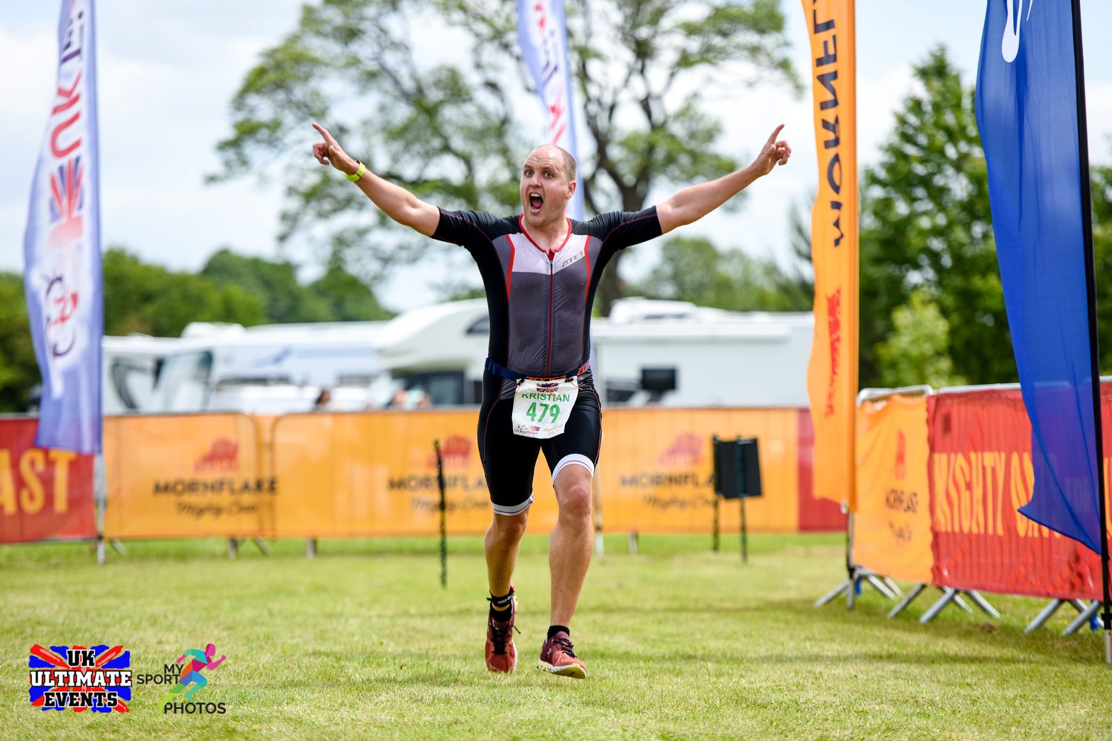 A man in a black and red triathlon suit joyfully crosses the finish line at an outdoor sports event. His arms are raised in celebration. He wears a race bib numbered 479. Banners and spectators are visible in the background.