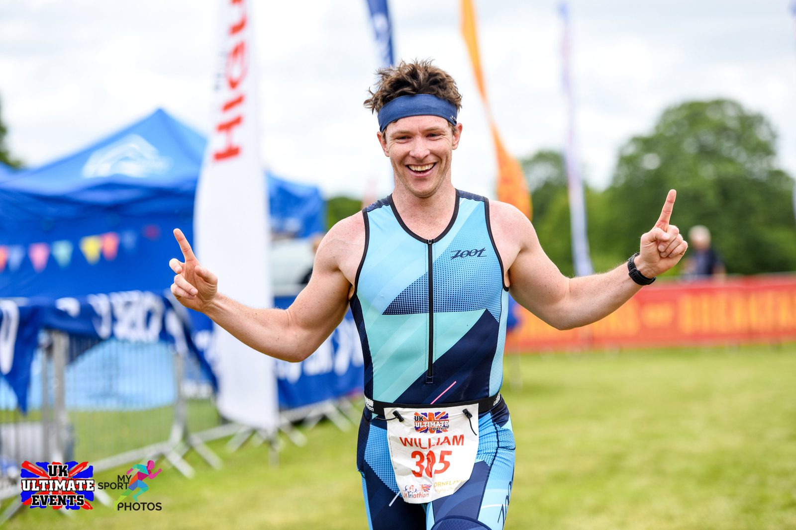 A male athlete, wearing a blue and black triathlon suit, raises both hands with index fingers pointed up, smiling as he crosses the finish line. He has a headband and a race bib with the name "William" and number "335." Banners and flags are visible in the background.