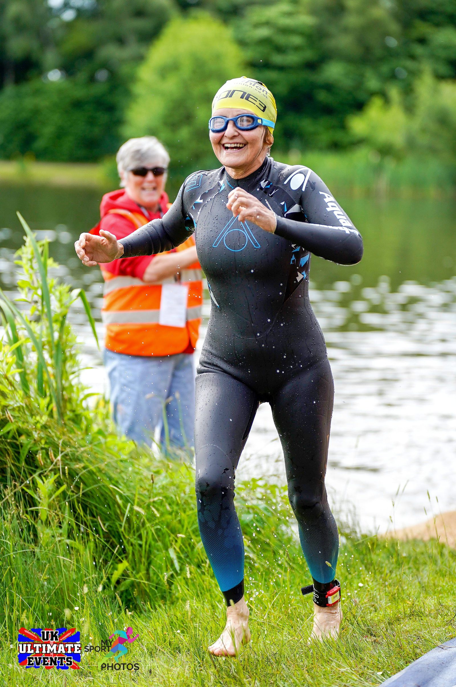 A swimmer in a wetsuit and swim cap, smiling and dripping wet, exits a lake during a triathlon event. Another person in a safety vest stands in the background near the water. The scene is outdoors with greenery and a lake in the background. Event logos are visible.