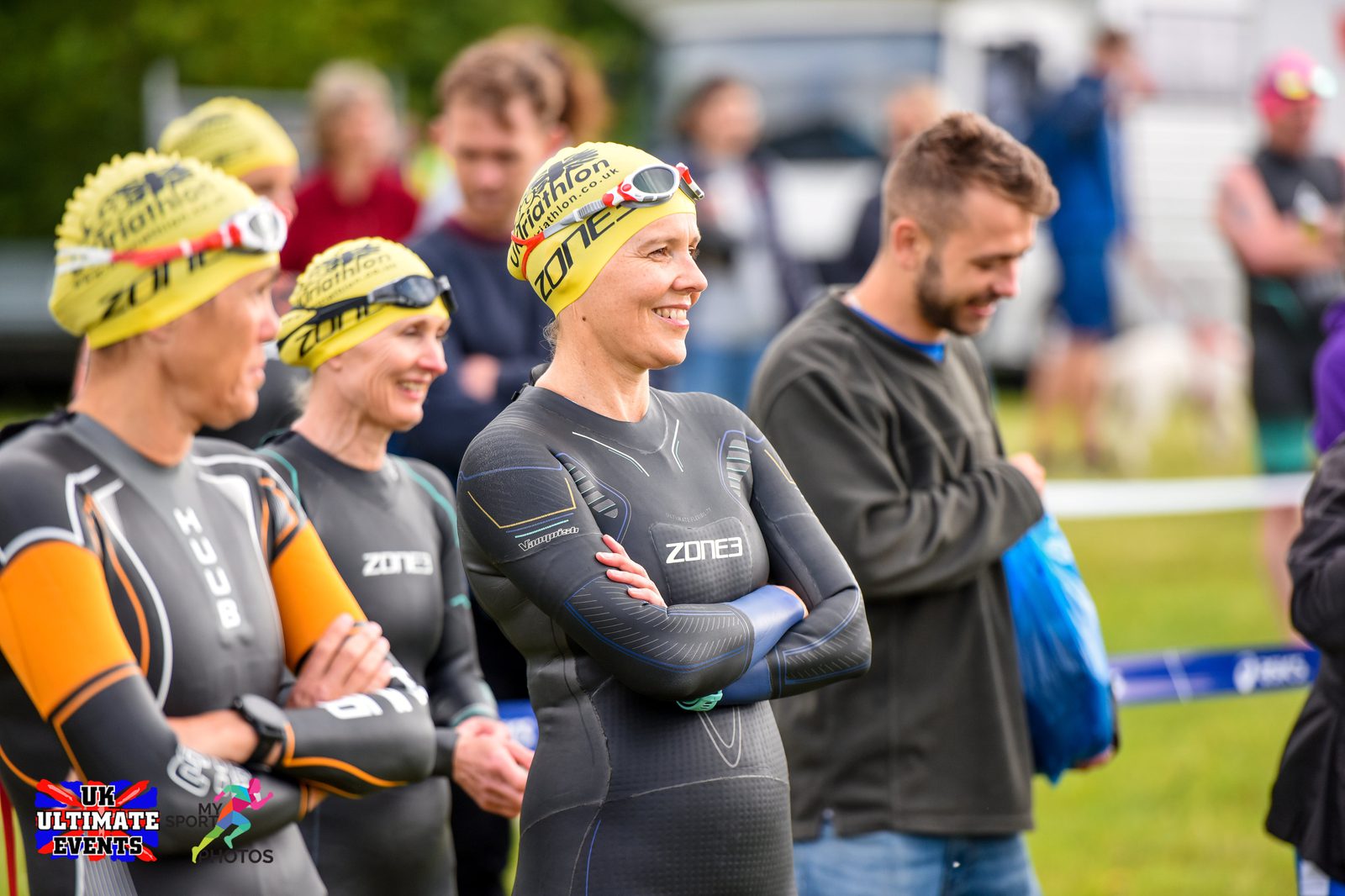 A group of athletes, wearing wetsuits and yellow swim caps, stand together at a triathlon event. They appear to be in good spirits, smiling and chatting. The background shows other participants and spectators, along with event banners and equipment.