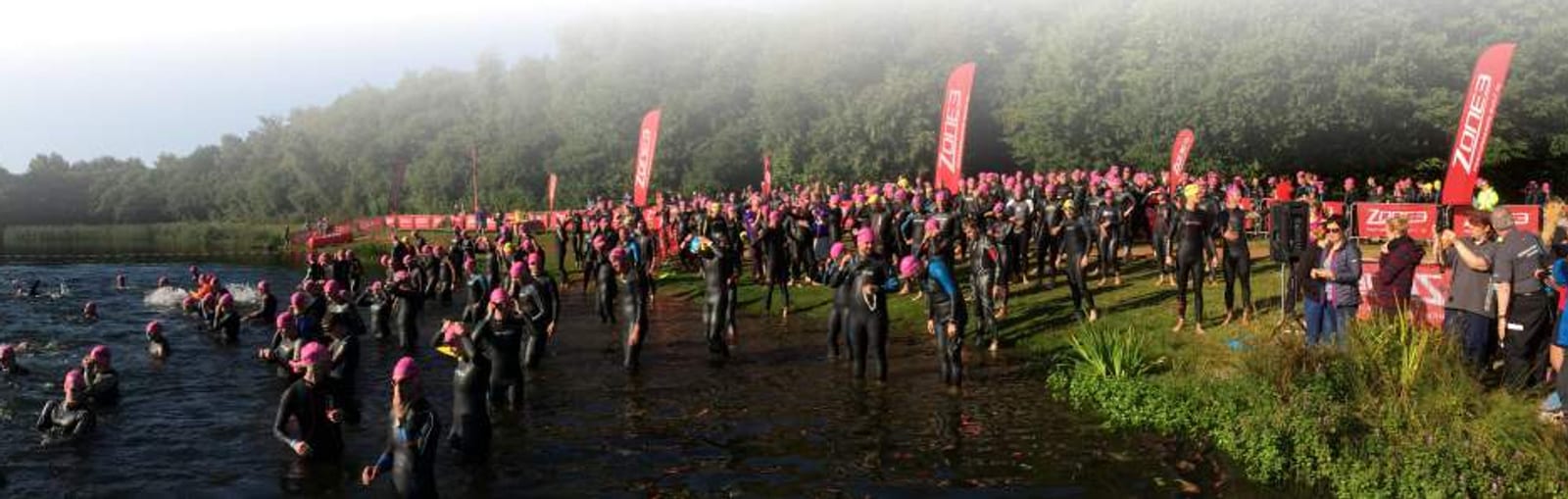 A large group of people in wetsuits and pink swim caps gather at the edge of a lake, preparing to swim. Red flags with logos are visible onshore, and trees with light fog in the background add to the scene. Some people are already in the water, while others stand on the bank.